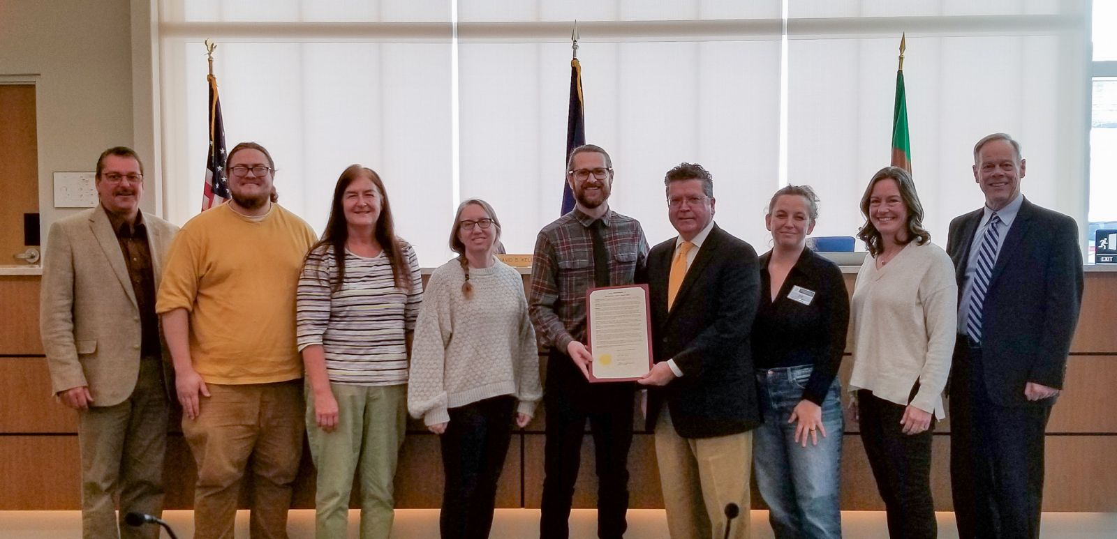 Group of eight people standing indoors, with one person holding a framed certificate. Flags are displayed in the background.