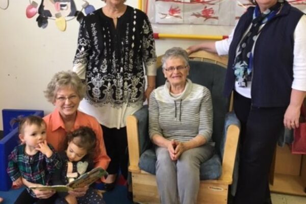 Four women and two children are in a room with colorful decorations. Two women are sitting and reading to the children, while two others stand behind them. A wall mural is visible in the background.