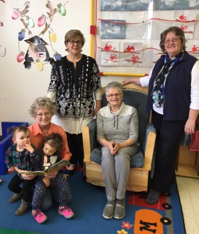 Four women and two children are in a room with colorful decorations. Two women are sitting and reading to the children, while two others stand behind them. A wall mural is visible in the background.