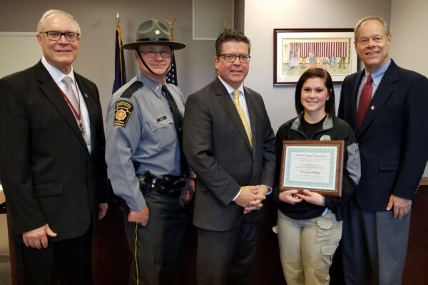 Five people stand indoors, one in a police uniform. A woman in uniform holds a framed certificate. They are positioned in front of a wall with a small American flag.