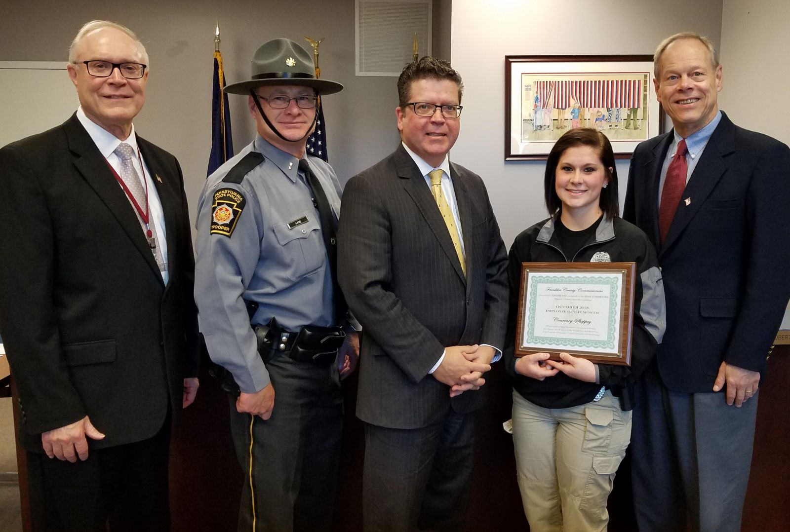 Five people stand indoors, one in a police uniform. A woman in uniform holds a framed certificate. They are positioned in front of a wall with a small American flag.