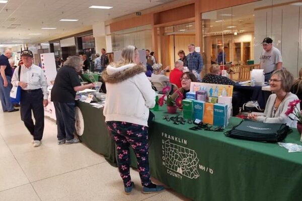 People visit various booths at a community fair inside a mall, with tables displaying informational materials and representatives engaging with attendees.