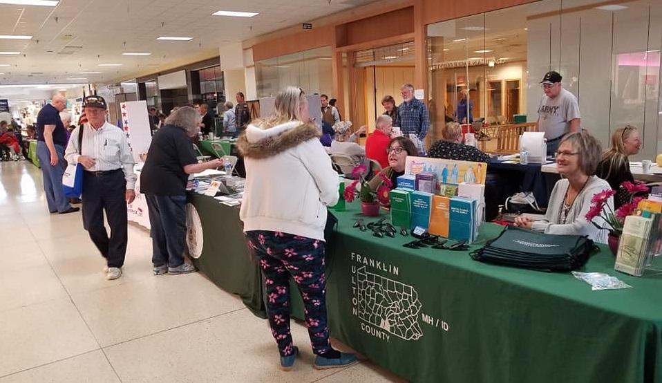 People visit various booths at a community fair inside a mall, with tables displaying informational materials and representatives engaging with attendees.