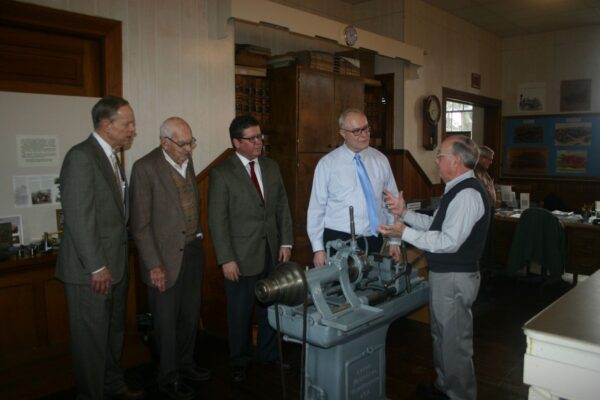 A group of five men in formal attire stand around an old machinery exhibit in a room full of historical memorabilia.