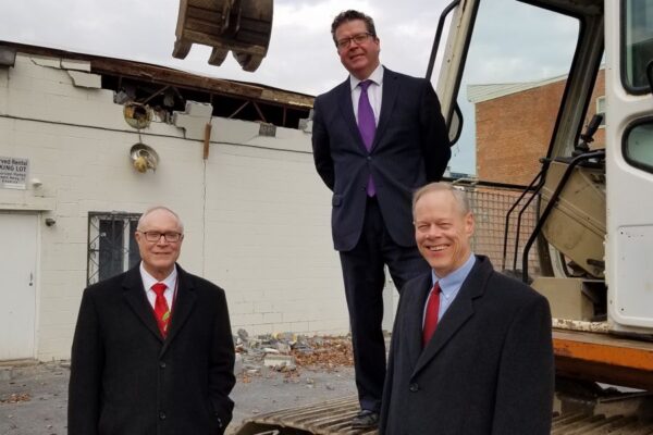 Three men in suits stand in front of a construction vehicle at a building demolition site. One man stands on the vehicle, while the other two stand on the ground.