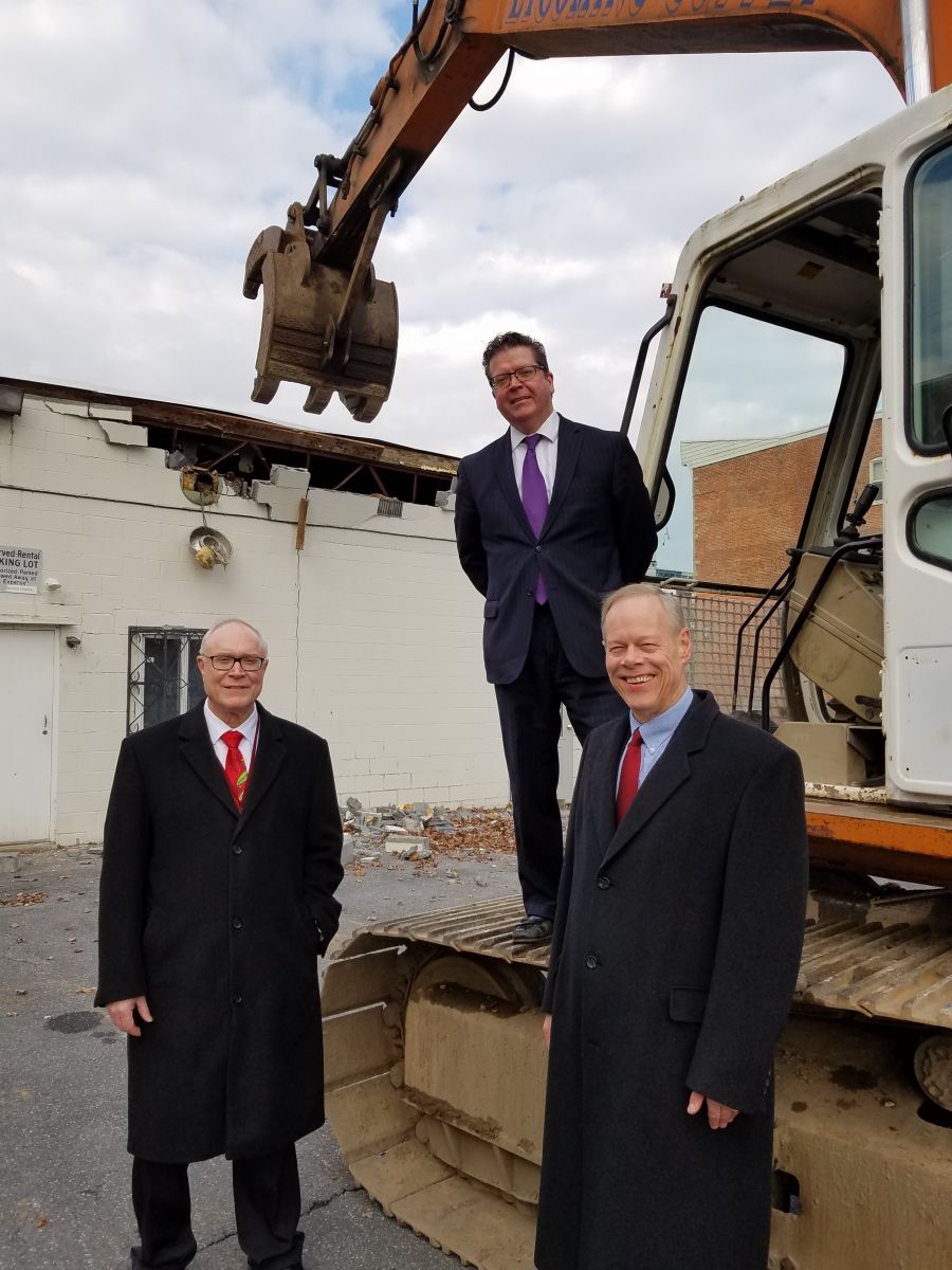 Three men in suits stand in front of a construction vehicle at a building demolition site. One man stands on the vehicle, while the other two stand on the ground.