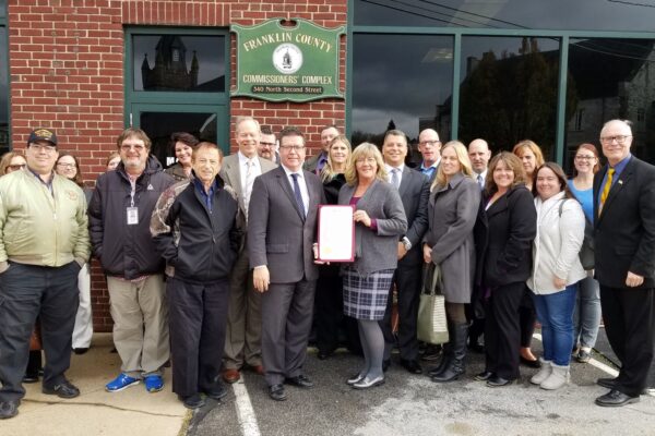 A group of people stands in front of Franklin County Commissioners' Complex. One person holds a framed document. They appear to be posing for a group photo.