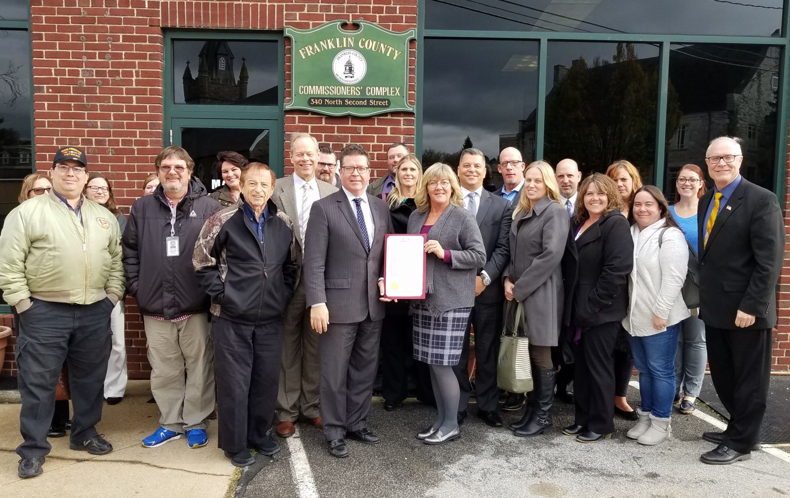 A group of people stands in front of Franklin County Commissioners' Complex. One person holds a framed document. They appear to be posing for a group photo.