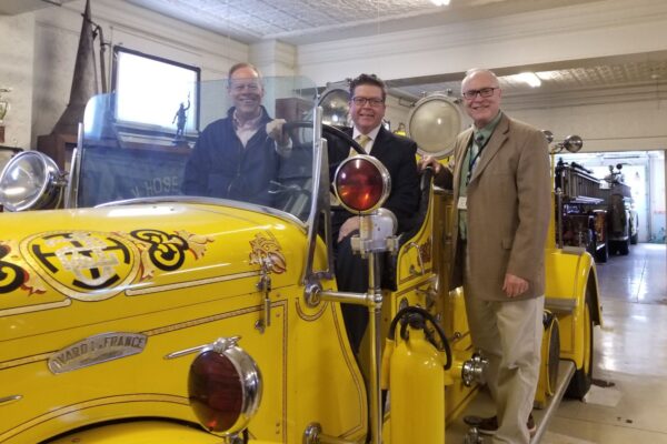 Three men stand beside a vintage yellow fire truck in a garage. One man is seated in the driver's seat, while the other two stand on either side. The garage has another fire truck in the background.