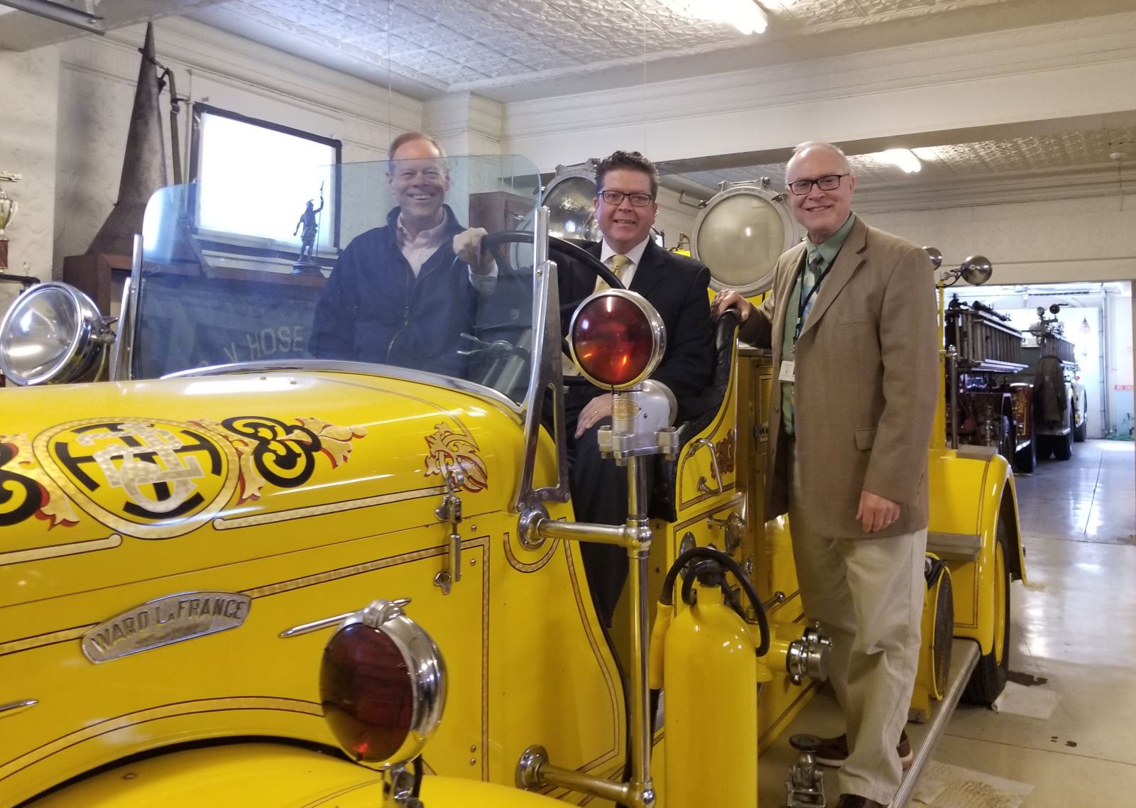 Three men stand beside a vintage yellow fire truck in a garage. One man is seated in the driver's seat, while the other two stand on either side. The garage has another fire truck in the background.