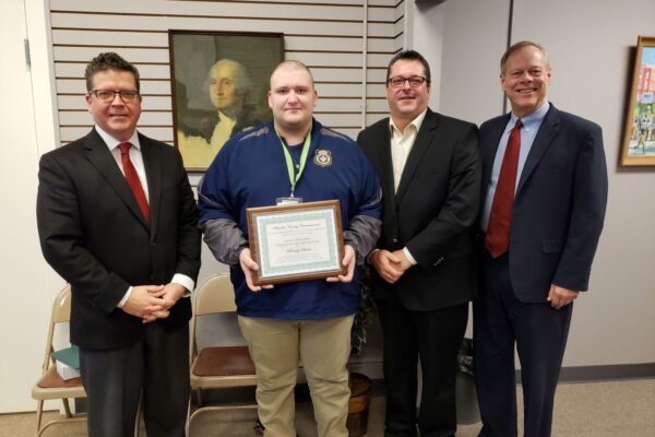 Four men are standing indoors, with one holding a certificate. There is a portrait on the wall behind them.