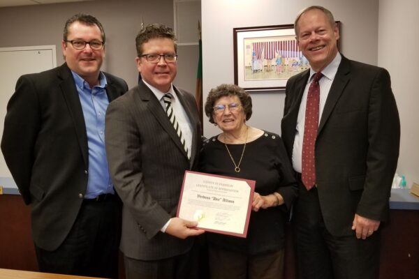 Four people standing together; an older woman holds a certificate, smiling. Three men in suits stand beside her in an office setting.