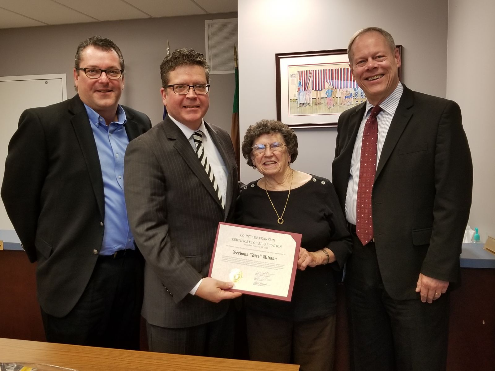 Four people standing together; an older woman holds a certificate, smiling. Three men in suits stand beside her in an office setting.