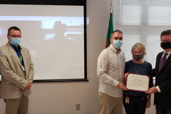 A group of people, some wearing masks, stand indoors. One person holds a certificate. A virtual participant is visible on a screen in the background.