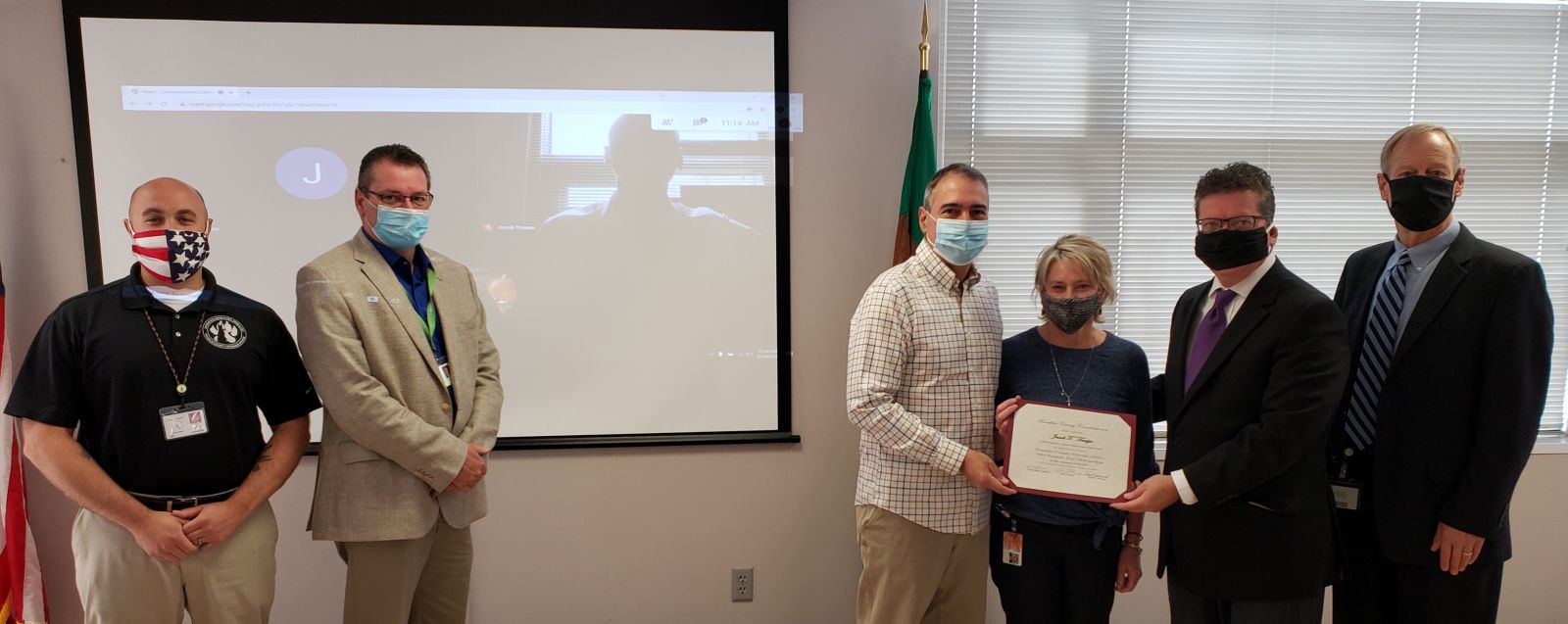 A group of people, some wearing masks, stand indoors. One person holds a certificate. A virtual participant is visible on a screen in the background.