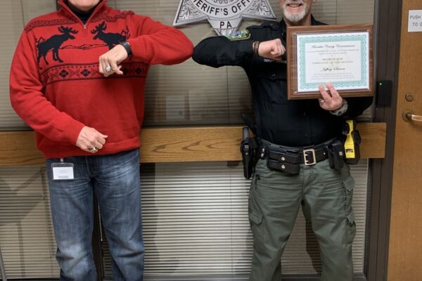 Two men touch elbows in front of a Franklin County Sheriff's Office sign. One wears a Christmas sweater; the other holds a certificate and wears a sheriff's uniform.