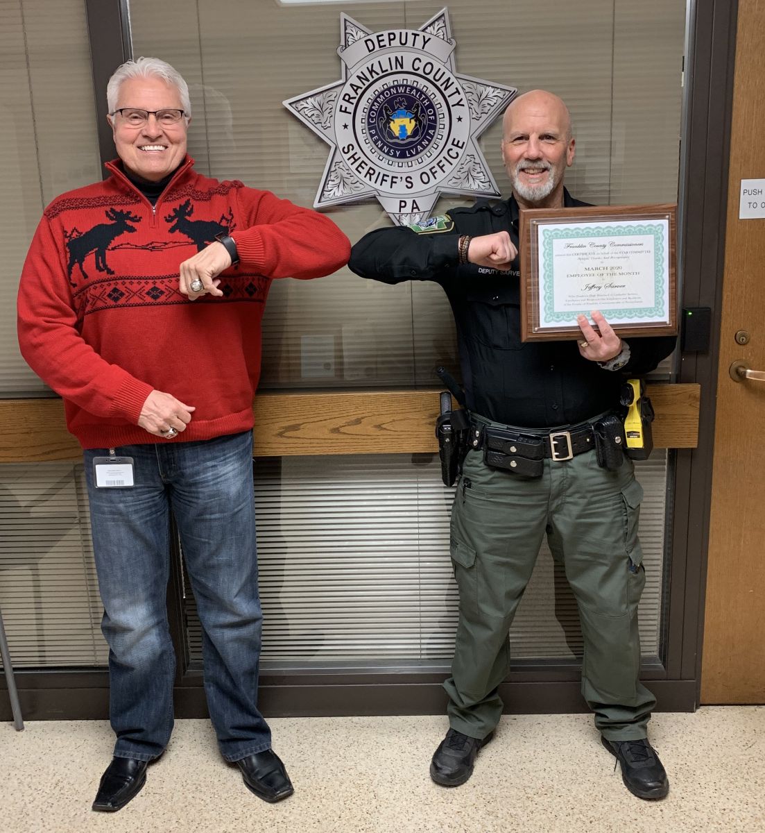 Two men touch elbows in front of a Franklin County Sheriff's Office sign. One wears a Christmas sweater; the other holds a certificate and wears a sheriff's uniform.