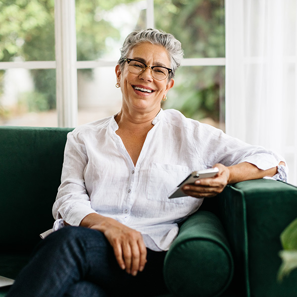 Happy elderly woman looking at the camera while sitting on a comfy couch with a smartphone.