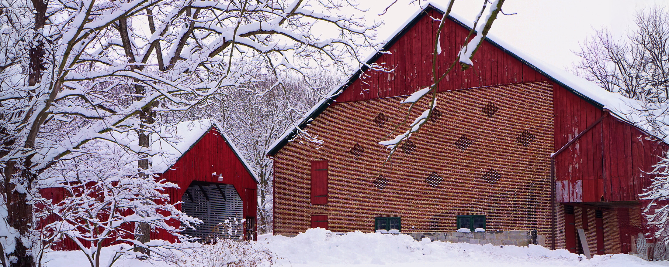 Red barn with brick facade and snow-covered trees and ground, under a cloudy winter sky.