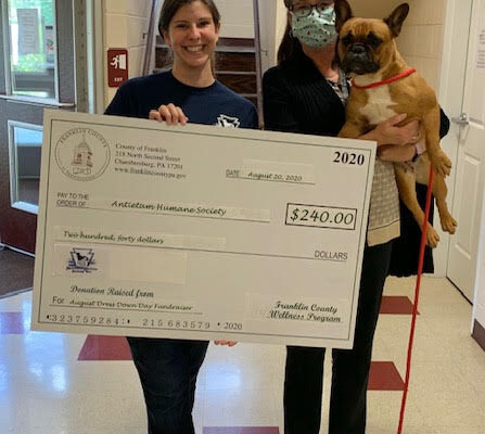 Two women stand indoors; one holds a large check for $240 addressed to the Animal Care Humane Society, and the other holds a small dog on a leash.