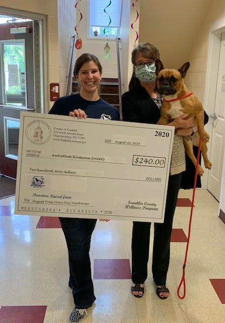 Two women stand indoors; one holds a large check for $240 addressed to the Animal Care Humane Society, and the other holds a small dog on a leash.