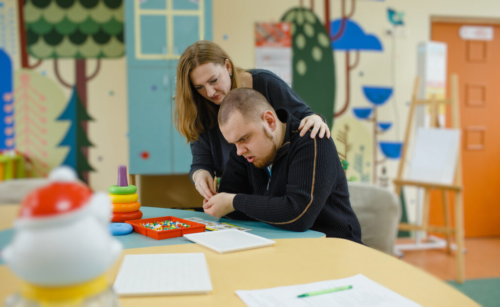 A woman assists a man with a puzzle in a colorful room, featuring a table with toys and educational materials.