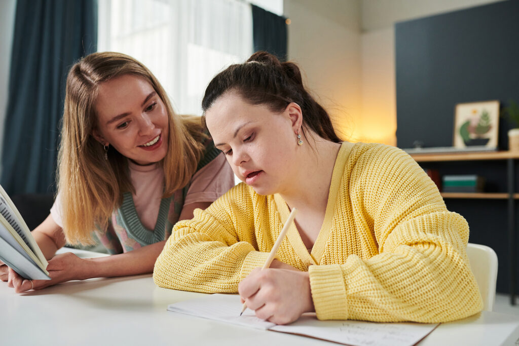 A woman in a yellow sweater writes in a notebook at a table, while another woman beside her smiles and looks on.