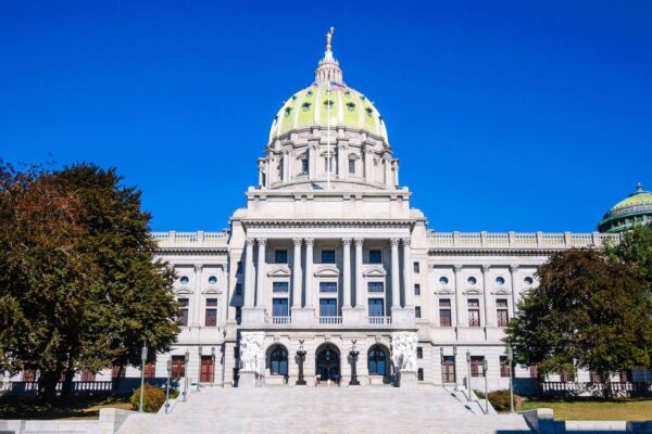 Front view of a grand building with a green-domed roof, white stone facade, steps leading up, and trees flanking each side under a clear blue sky.