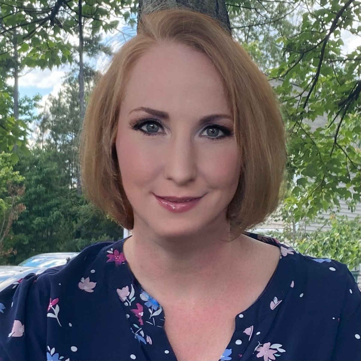 Woman with short auburn hair wearing a navy floral blouse, standing outdoors against a backdrop of trees.