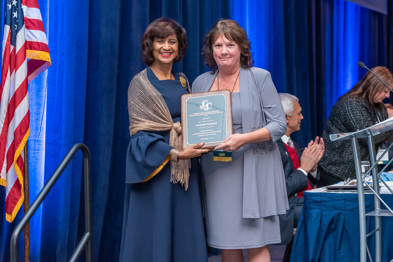 Two women stand side by side, one holding a plaque. They are on a stage with blue curtains and an American flag in the background.