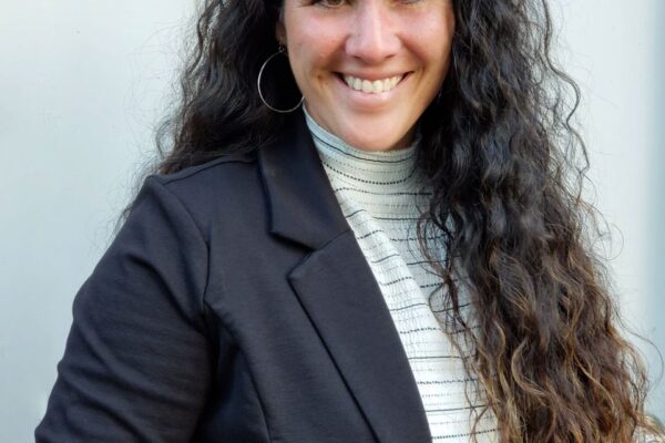 A person with long, curly hair is smiling while wearing a black blazer and striped shirt.