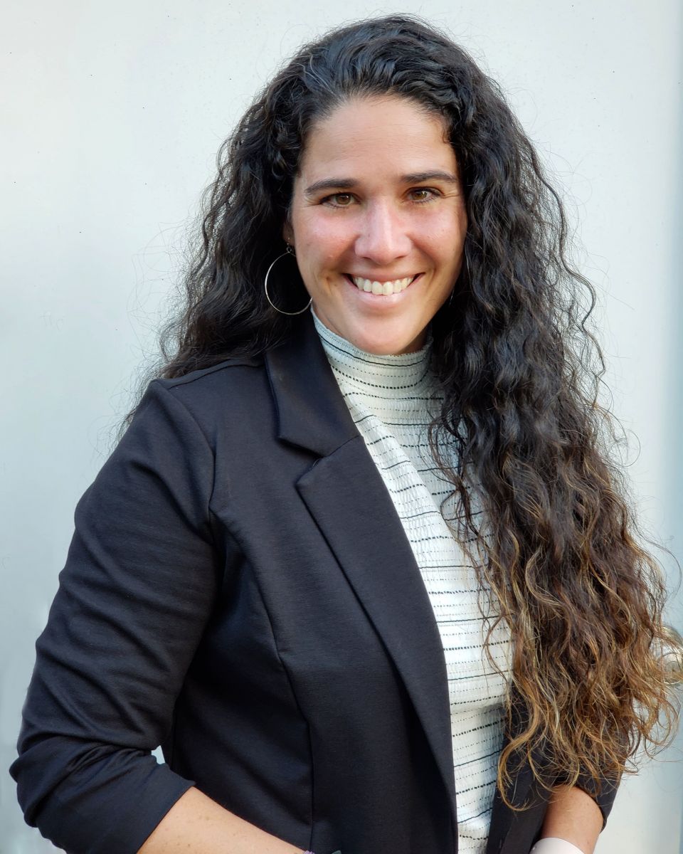 A person with long, curly hair is smiling while wearing a black blazer and striped shirt.