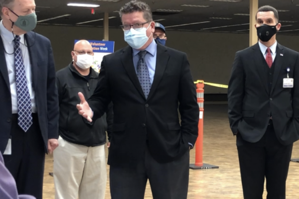 A group of men in suits, wearing face masks, stand indoors. One man in the front center is speaking, while others listen.