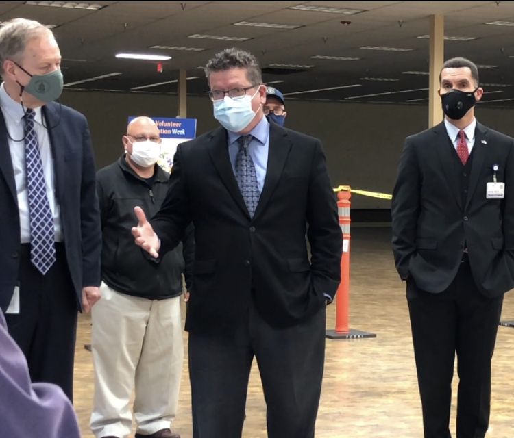 A group of men in suits, wearing face masks, stand indoors. One man in the front center is speaking, while others listen.