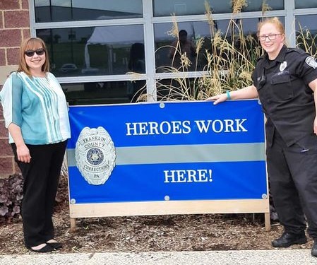 Two people stand beside a blue sign that reads "Heroes Work Here!" outside a building. One person is in uniform, the other in casual attire.