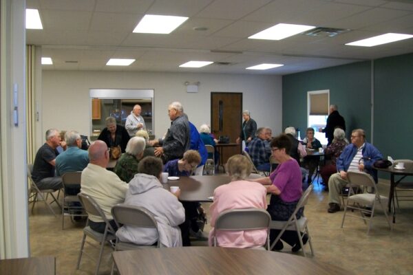 A group of older adults are gathered in a community room, sitting and standing around tables, engaged in conversation.