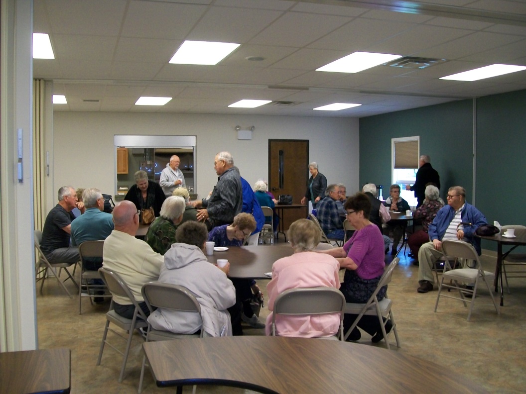 A group of older adults are gathered in a community room, sitting and standing around tables, engaged in conversation.