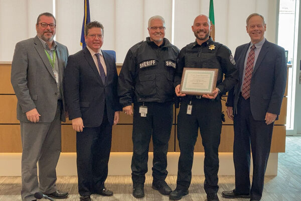 Five men standing in a row; two wearing sheriff uniforms, one holds a framed certificate.