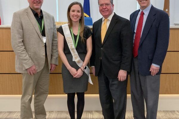 Four people standing together; the woman is wearing a sash, and they are all dressed in business attire, posing indoors in front of a wooden panel with flags.