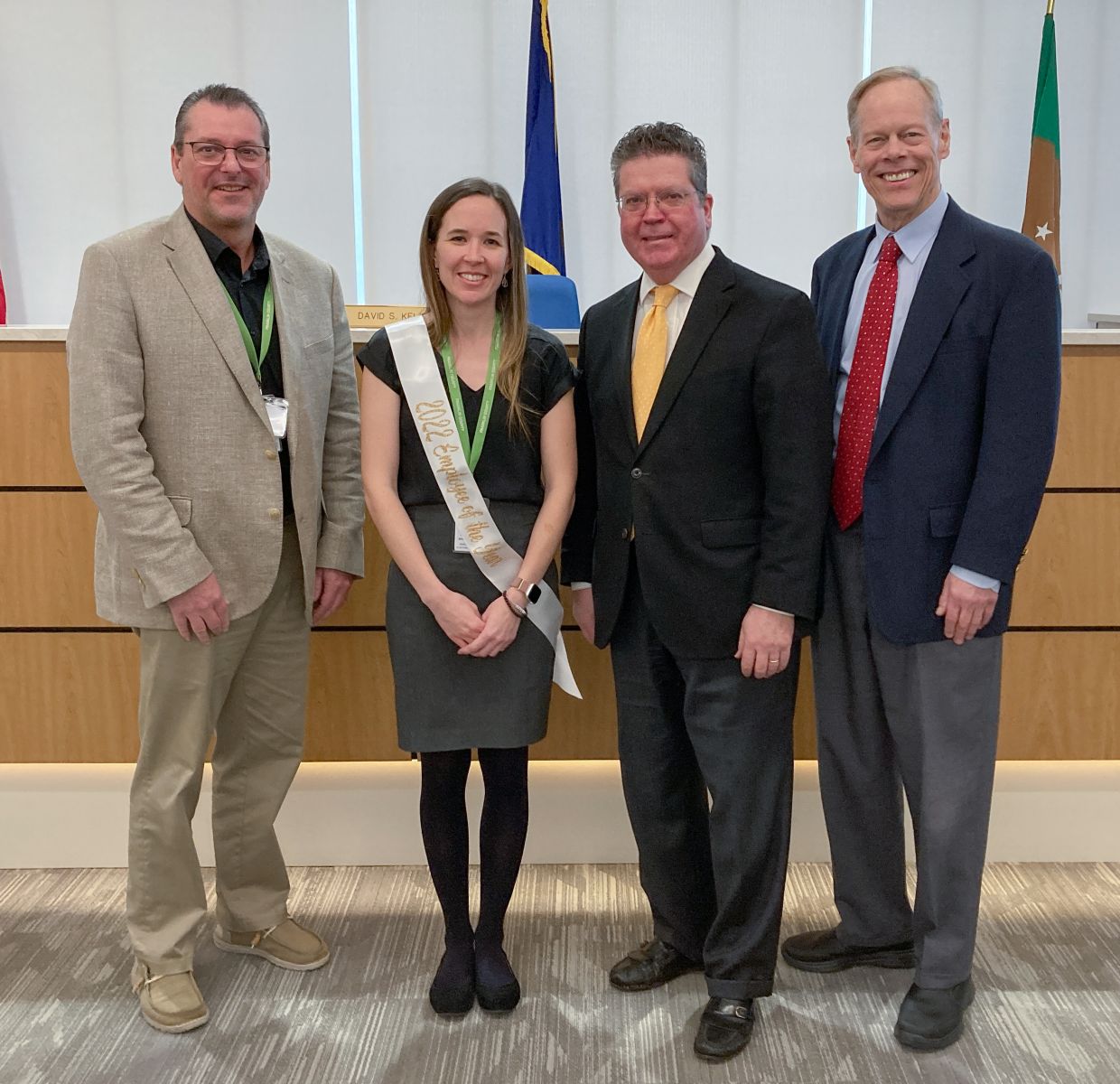 Four people standing together; the woman is wearing a sash, and they are all dressed in business attire, posing indoors in front of a wooden panel with flags.