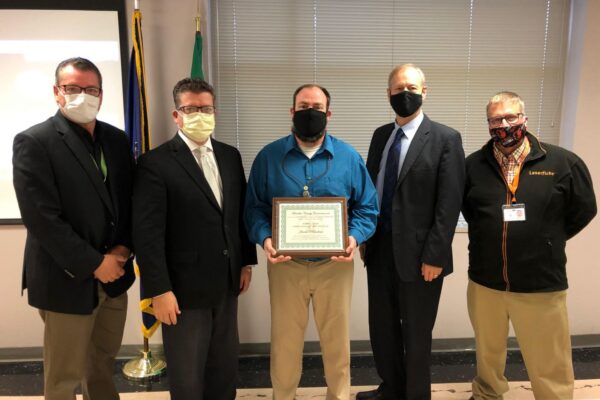 Five men in business attire and masks stand in a line; the middle person holds a certificate. Flags are partially visible in the background.