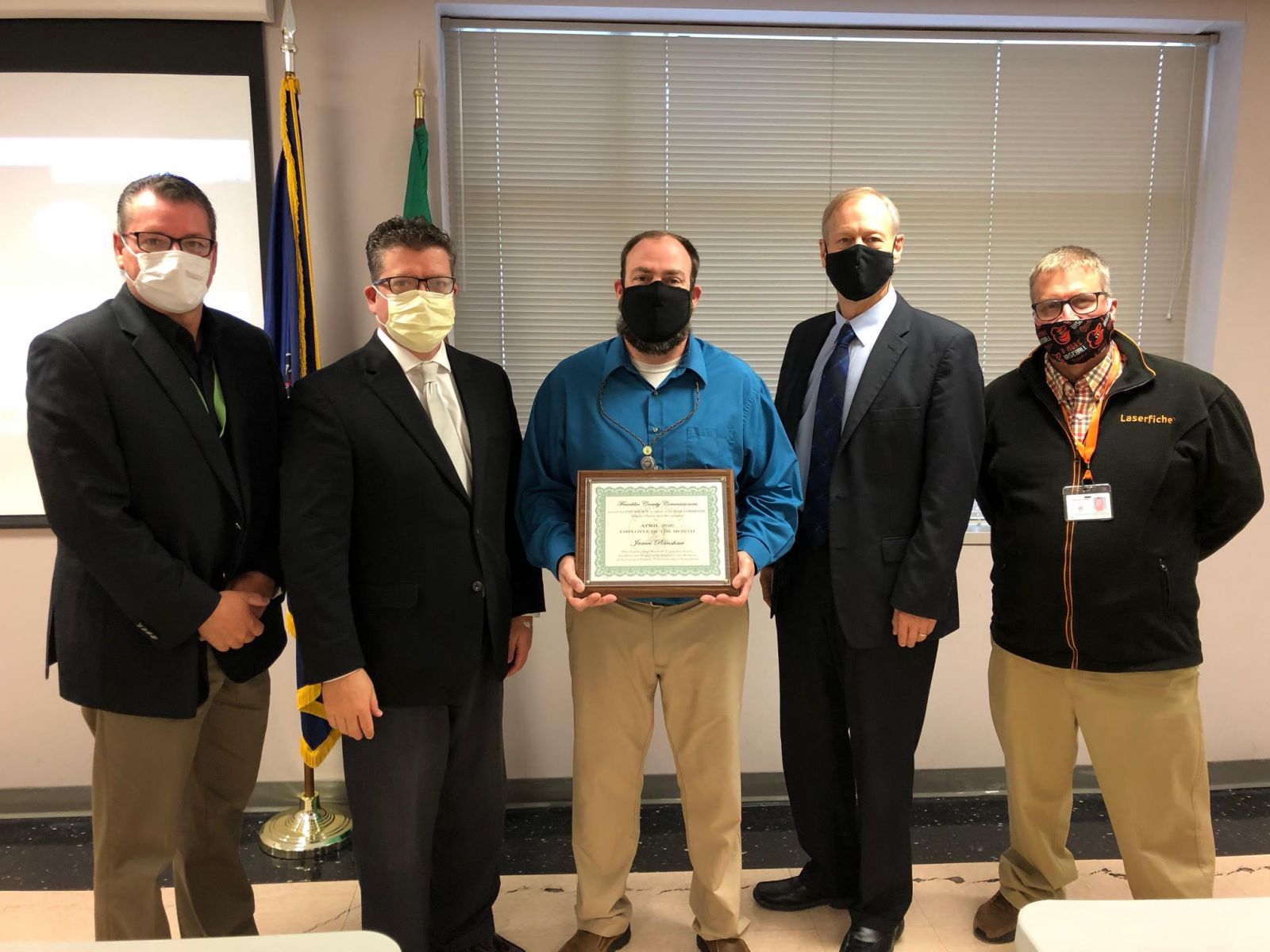 Five men in business attire and masks stand in a line; the middle person holds a certificate. Flags are partially visible in the background.