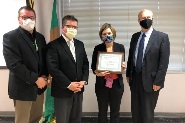 Four people stand together, with the person second from the right holding a framed certificate. They are all wearing masks. A flag and a window with blinds are visible in the background.