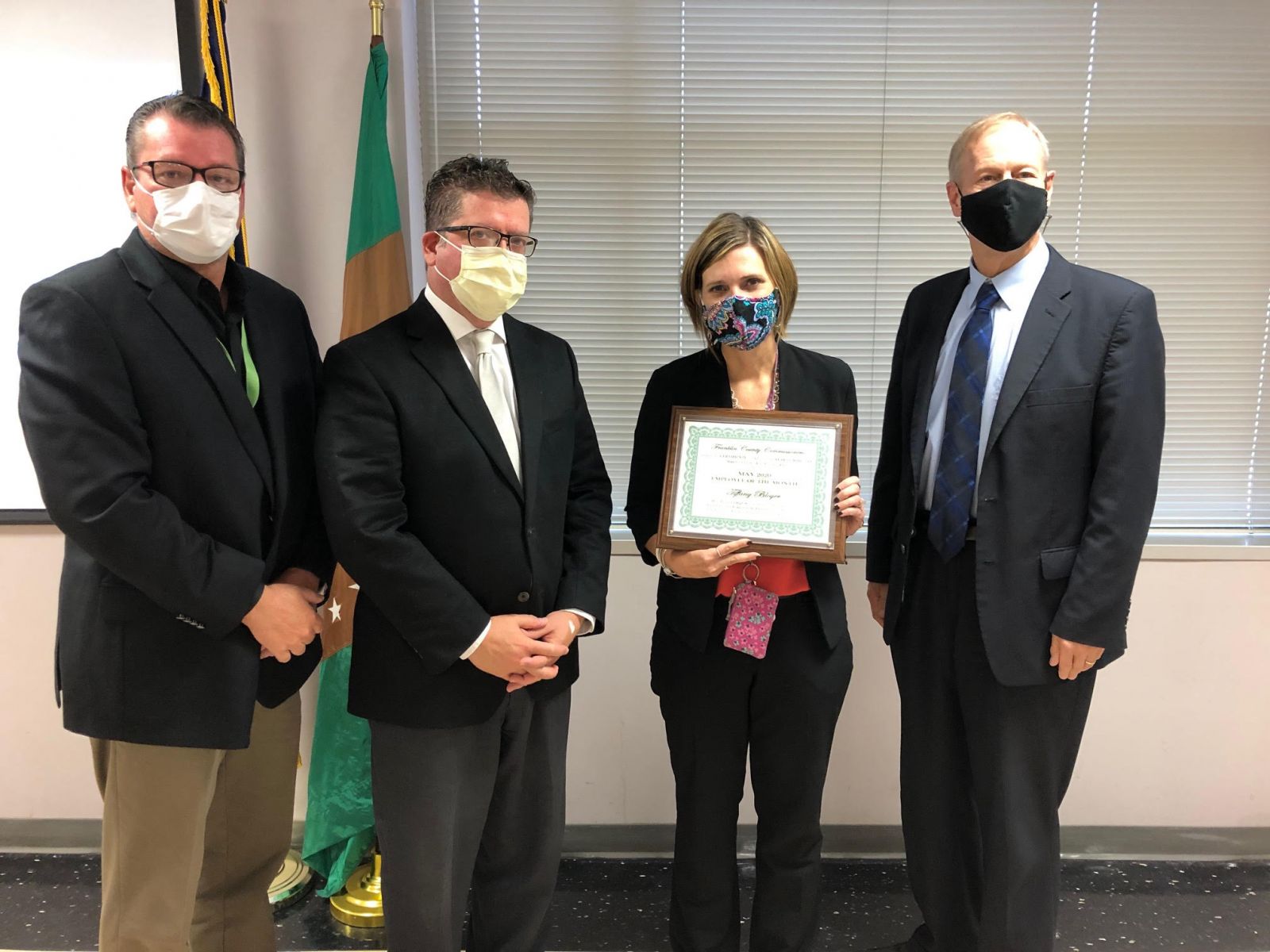 Four people stand together, with the person second from the right holding a framed certificate. They are all wearing masks. A flag and a window with blinds are visible in the background.