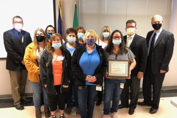 A group of people wearing masks pose indoors. One person holds a framed certificate.