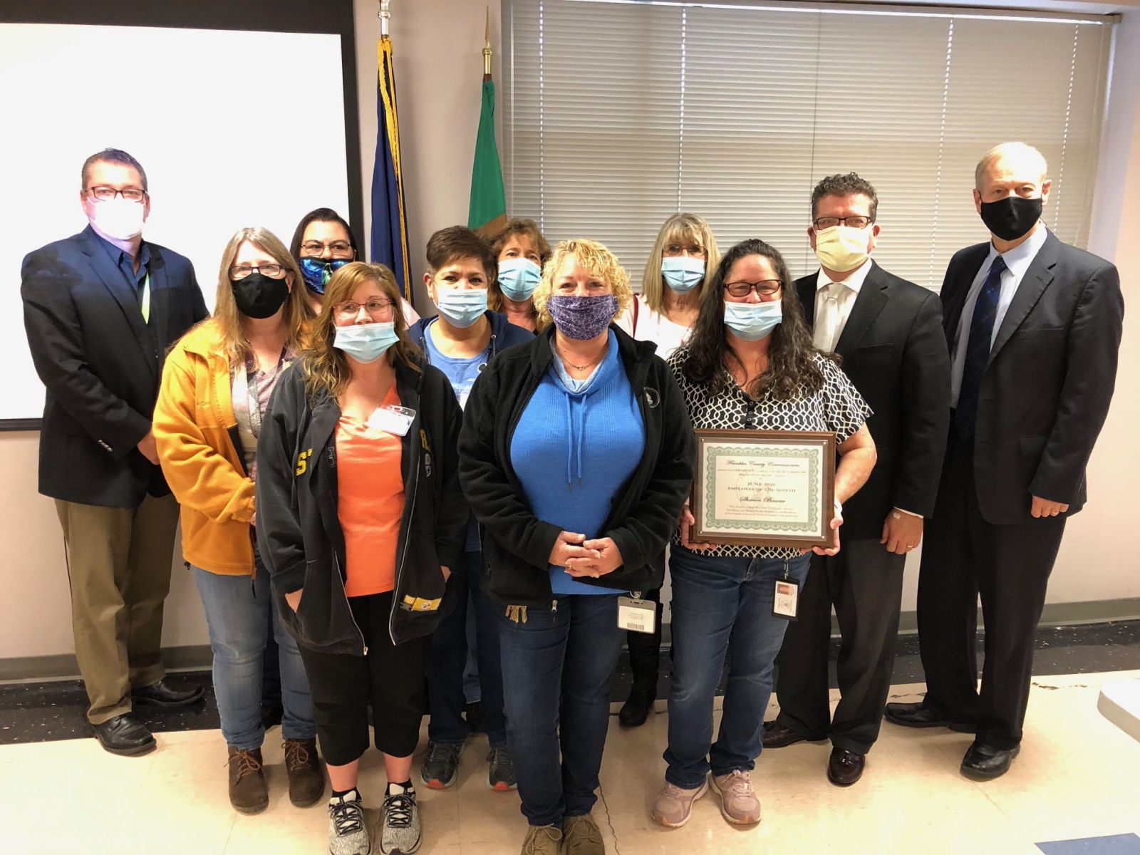 A group of people wearing masks pose indoors. One person holds a framed certificate.