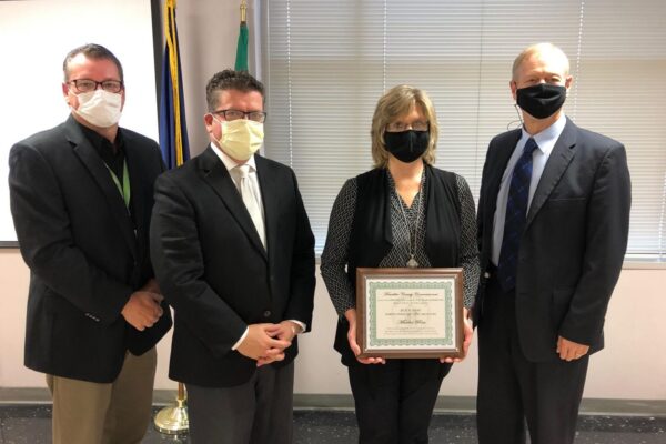 Four people wearing masks, three men in suits on either side of a woman holding a framed certificate, stand in a room with flags and blinds in the background.