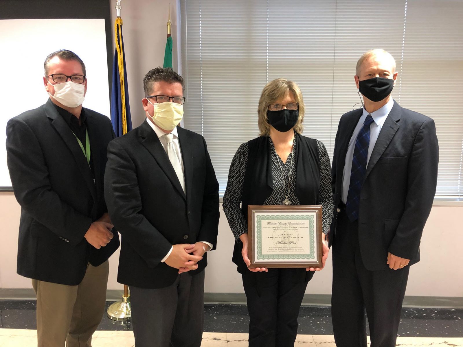 Four people wearing masks, three men in suits on either side of a woman holding a framed certificate, stand in a room with flags and blinds in the background.