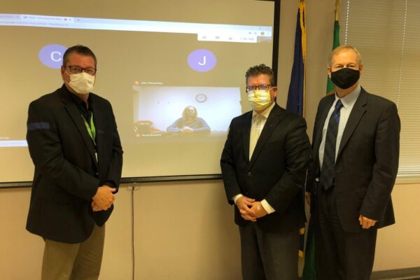 Three men in suits and face masks stand indoors in front of a projected screen showing a video call with two participants.