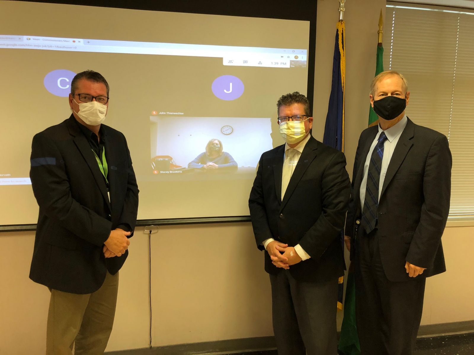 Three men in suits and face masks stand indoors in front of a projected screen showing a video call with two participants.